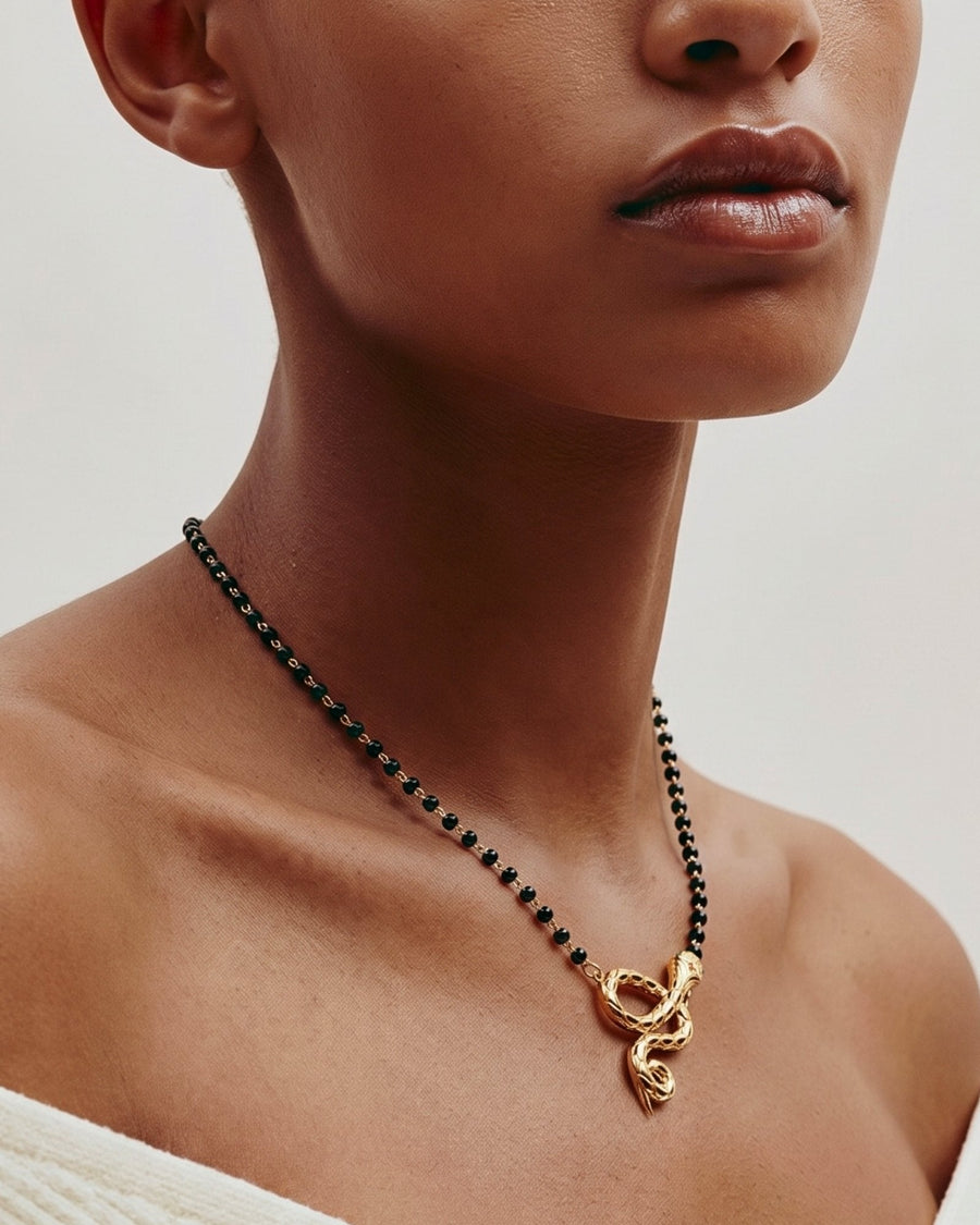 The Ammama 18K gold twin serpent necklace with emerald eyes on a black-beaded chain